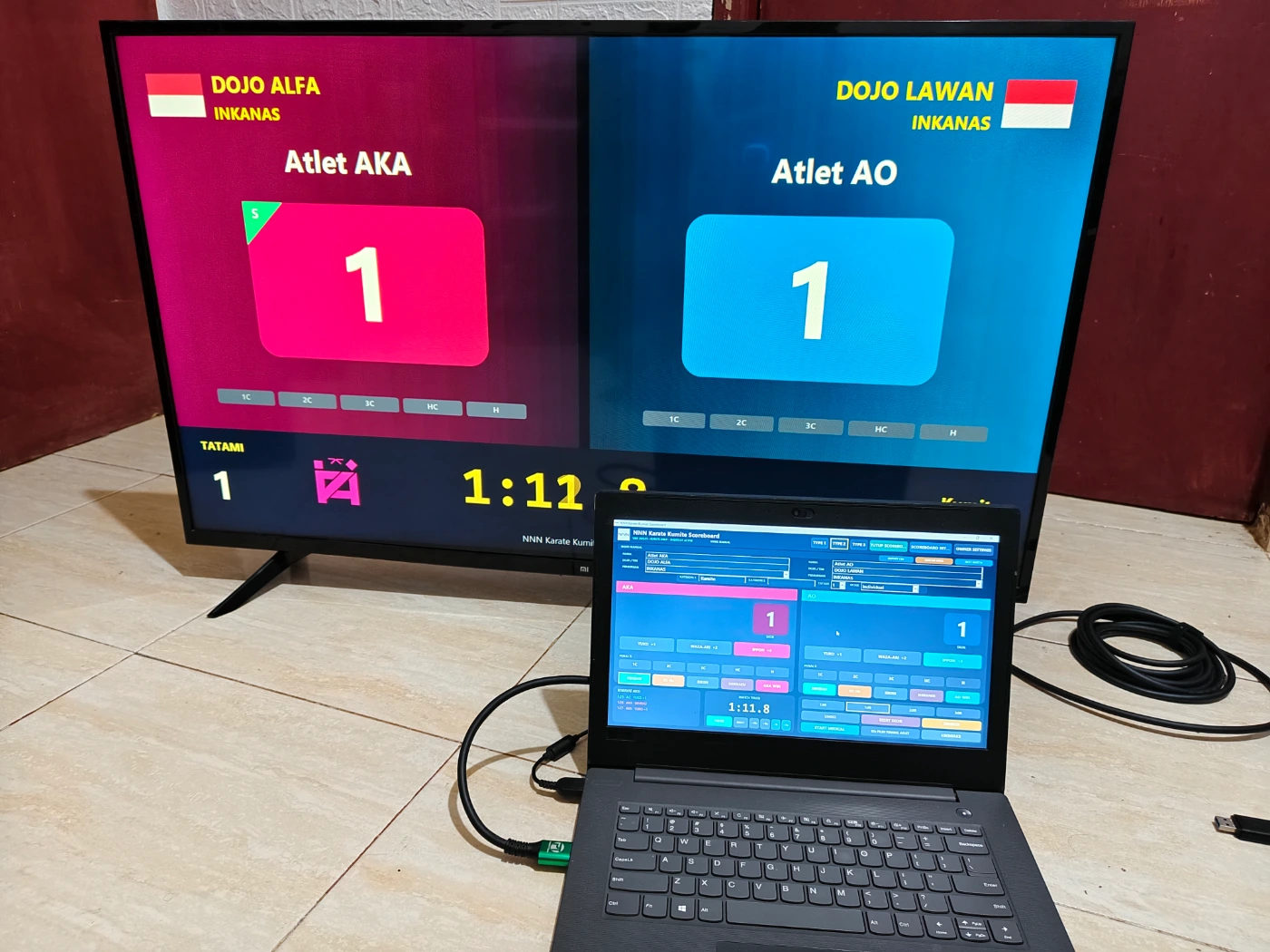 Operator menggunakan aplikasi papan skor Karate Kumite pada laptop Windows dengan tampilan skor di TV